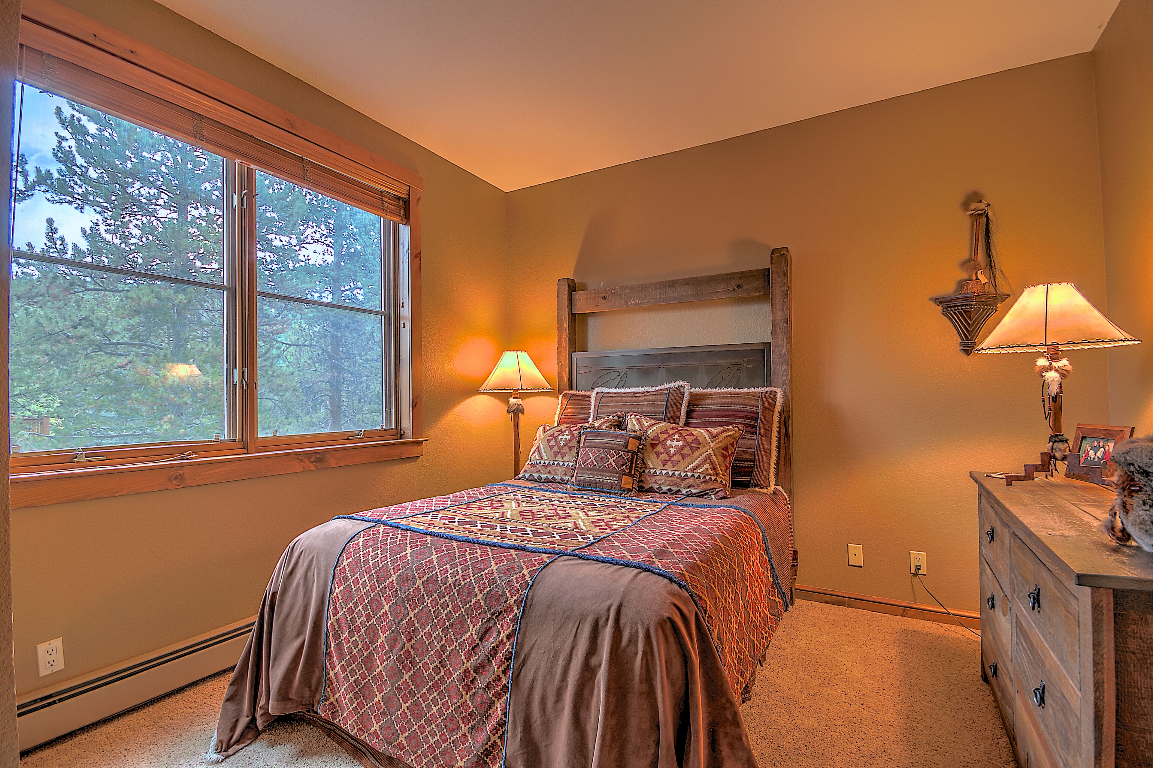 A bedroom with a large window, a bed with patterned bedding, two bedside lamps, and a dresser with a mirror and picture frame.