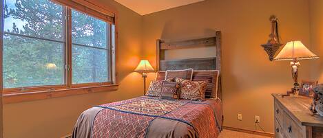 A bedroom with a large window, a bed with patterned bedding, two bedside lamps, and a dresser with a mirror and picture frame.