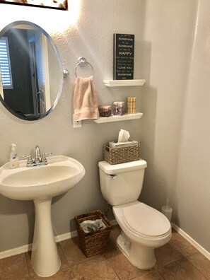 A bathroom with a sink, toilet, and a mirror. There is a towel hanging on the wall, a shelf with toiletries, and a basket on the floor.