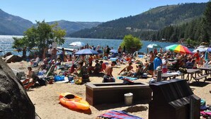 A crowded beach with people sunbathing and swimming, surrounded by mountains and clear blue water.