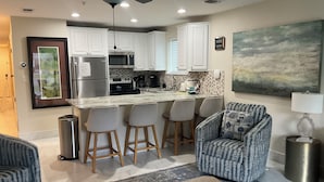 Una cocina moderna con armarios blancos, una isla central con taburetes y un comedor con sillas estampadas, además de un cuadro abstracto grande en la pared.