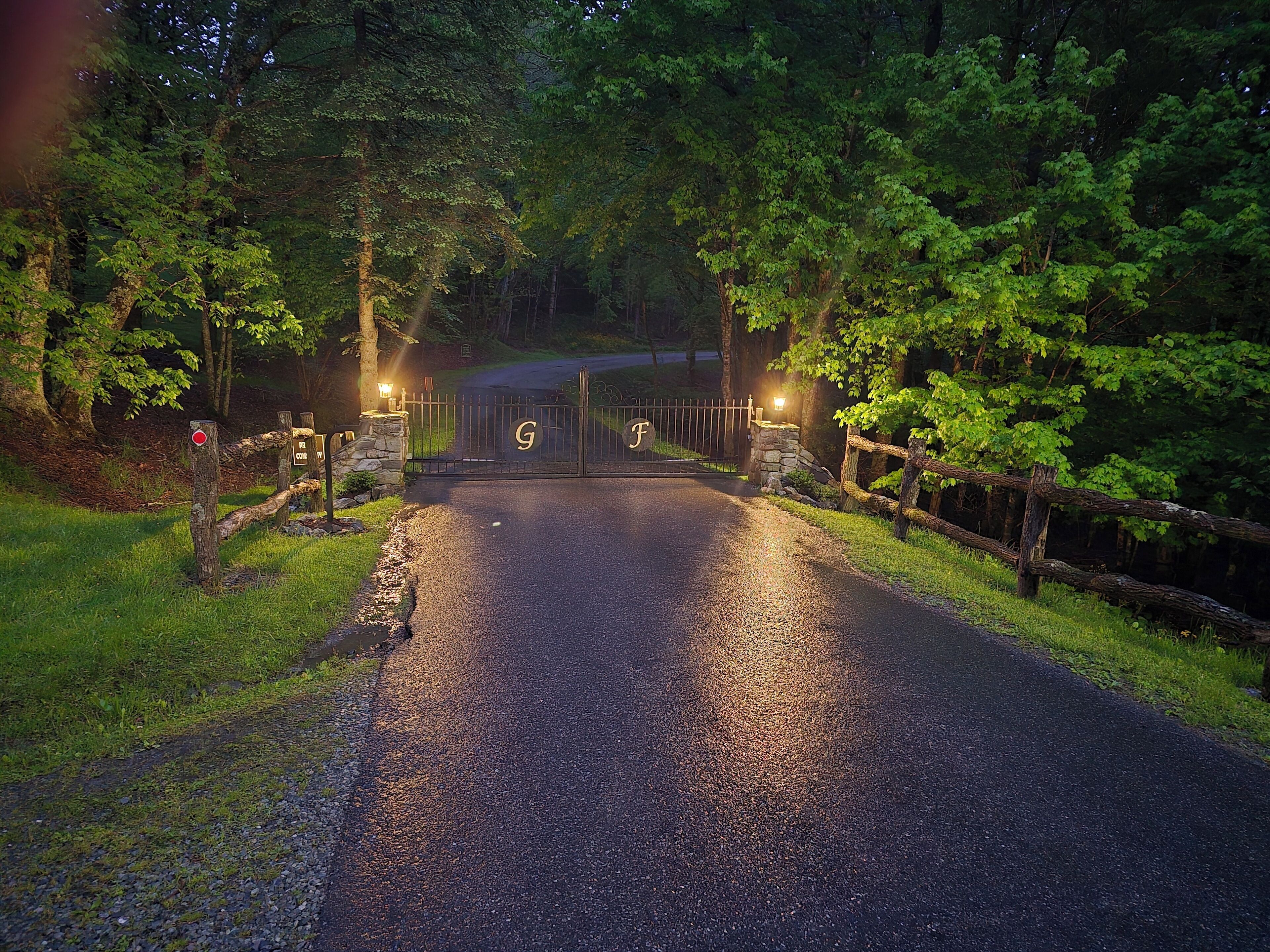 A gated driveway leading into a wooded area with a stone wall and a wooden fence.