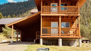 A two-story wooden cabin with a balcony, a covered porch, and a mountain backdrop. - 12 miles to Yellowstone National Park, hot tub, fire pit, 3 bedrooms 3 bathrooms (Island Park)