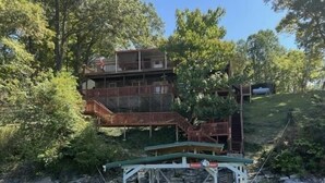 Un bote amarrado con una estructura cubierta, un edificio de varios niveles con una escalera de madera, y un paisaje natural con árboles y una colina.