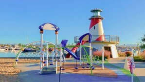 Ein Spielplatz mit Rutschen und Klettergeräten, ein Leuchtturm und im Hintergrund ein Yachthafen.
