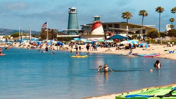 Ein Strand mit Leuchtturm, Sonnenschirmen und Menschen beim Schwimmen und Entspannen.