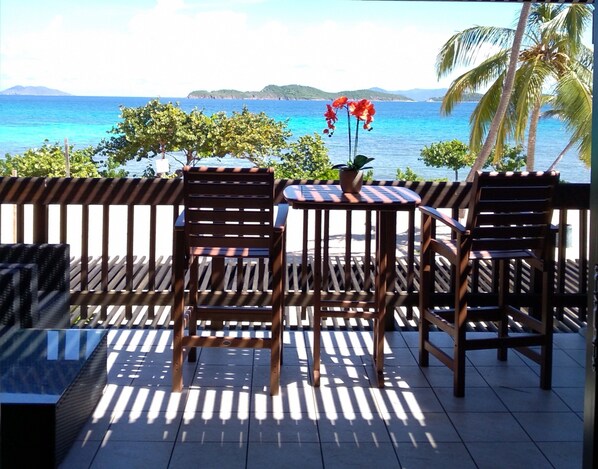 A wooden deck with two chairs and a table, overlooking a turquoise sea with distant hills.