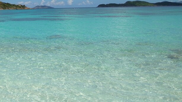 A clear turquoise sea with visible seabed, a distant shoreline with vegetation, and a partly cloudy sky. - IMMACULATE! WASTE NO TIME GETTING TO THE BEACH! (St. Thomas)