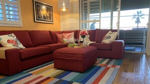 Un salon avec un canapé d’angle rouge, un tapis rayé aux couleurs vives et une grande fenêtre avec des stores.