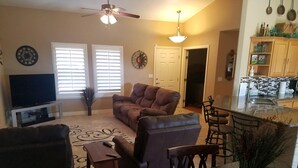 A living room with a brown leather sofa, a wooden coffee table, and a TV on a stand. There is a dining area with a table and chairs, and a kitchen visible in the background.