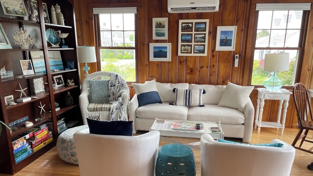 Una sala de estar acogedora con un sofá blanco, dos sillones, una mesita de centro y una estantería llena de libros y objetos decorativos.