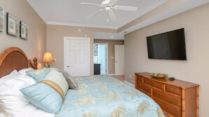 Un dormitorio con cama, cómoda, ventilador de techo, televisor y puerta que da a otra habitación.