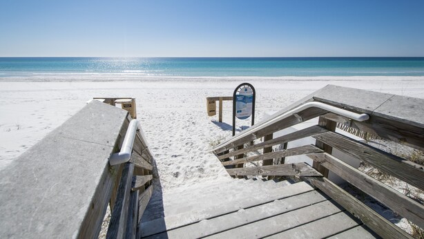 Ein Holzsteg führt zu einem Strand, an dem ein Schild steht.