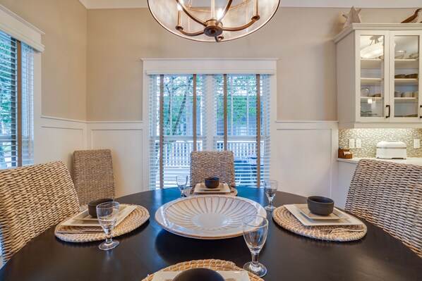 A dining area with a dark wooden table set for four, surrounded by wicker chairs, a large glass bowl centerpiece, and a view of a balcony through the window.