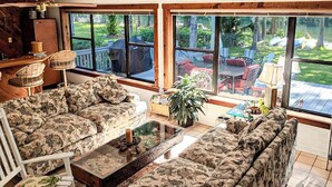 Un salon avec deux canapés à motifs, une table basse et un fauteuil à bascule. De grandes fenêtres offrent une vue sur le jardin.