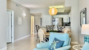 Un salon avec un fauteuil bleu clair, un coin repas avec une table et des chaises, et une cuisine à l’arrière-plan.