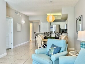 A living room with a light blue armchair, a dining area with a table and chairs, and a kitchen in the background. - Beautiful 6th Floor New LR furniture, 2BR/2BACondo- 2 King BR, 2 BA , BUNK ROOM! (Panama City Beach)