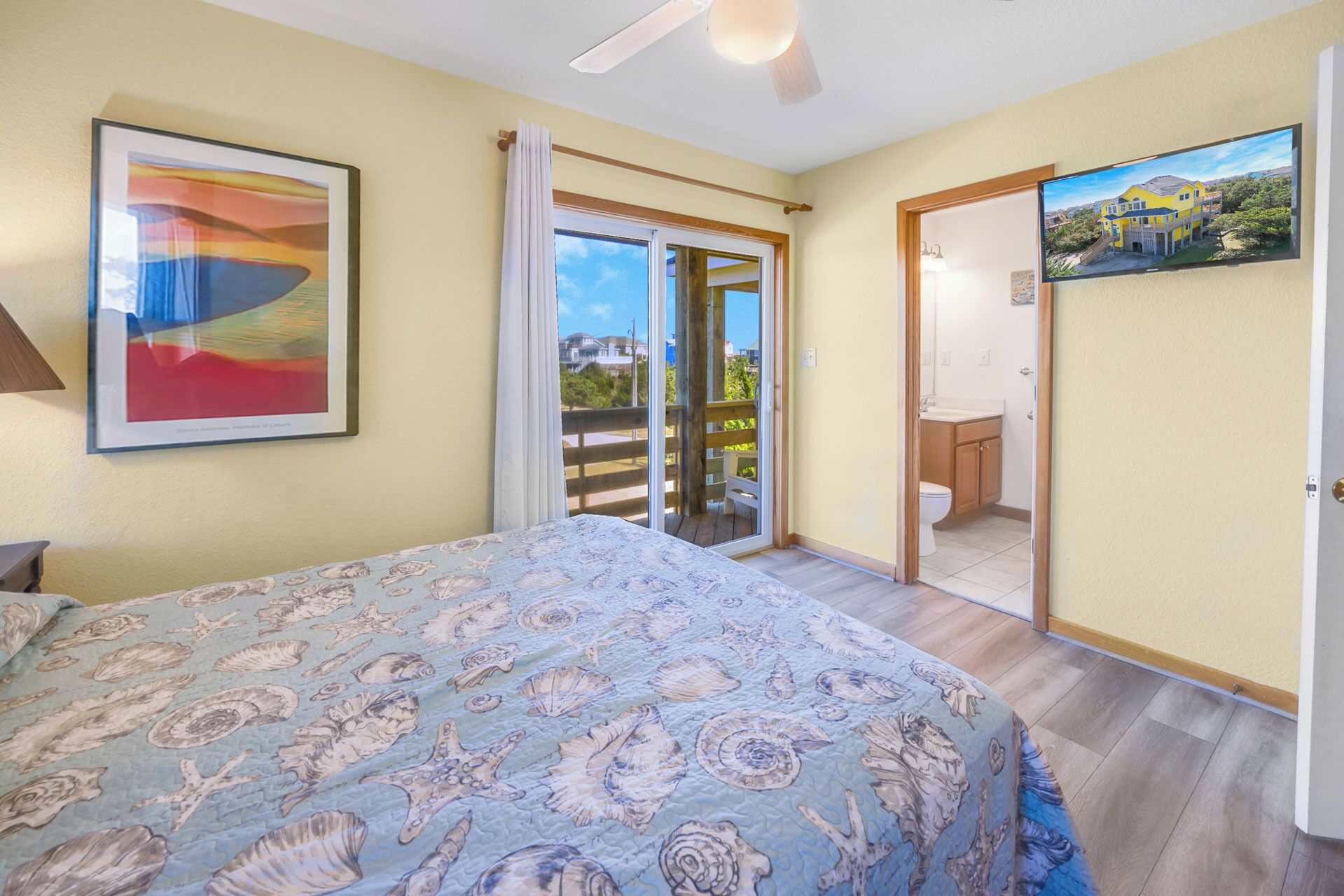 Quarto com cama, pintura, porta de vidro deslizante para uma varanda e banheiro visível através da porta.