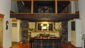 A spacious dining area with a high ceiling and exposed wooden beams, a kitchen with stone accents, and a balcony with a railing.