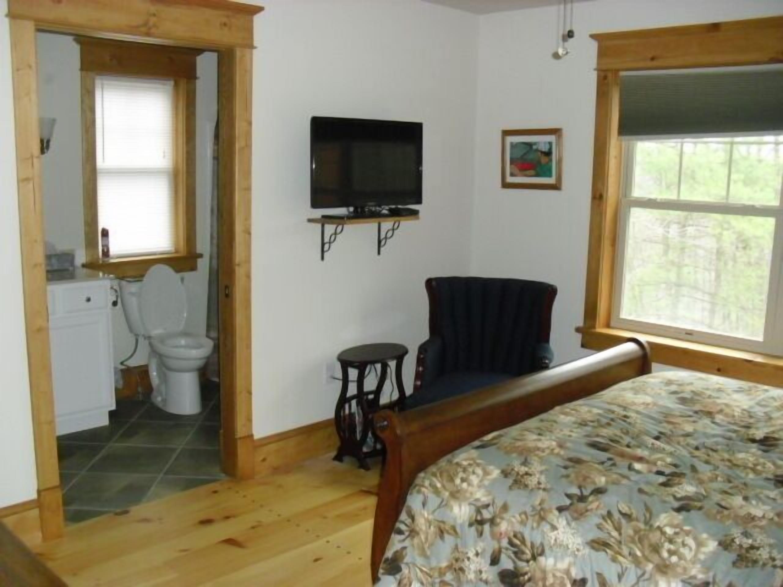 Ein Schlafzimmer mit einem hölzernen Bett, einem an der Wand befestigten Fernseher, einem Sessel und einem Fenster mit Blick auf Bäume.