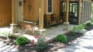 A wooden cabin with a porch, stone pillars, and a dining area with chairs and a table.