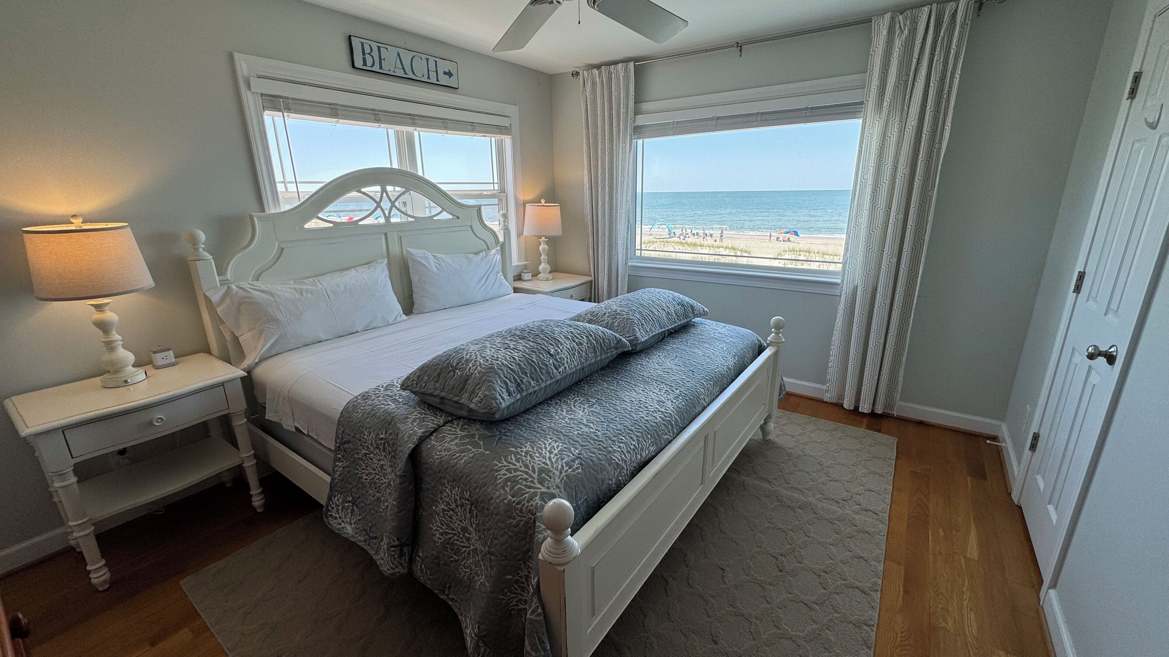 A bedroom with a bed, nightstands, a ceiling fan, and a view of the beach.