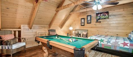 A cozy room with a pool table, wooden ceiling beams, a ceiling fan, and a television on the wall.