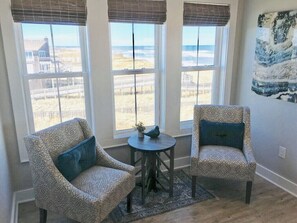Ein Zimmer mit zwei gemusterten Sesseln, einem kleinen runden Tisch mit einer Dekoration sowie Blick auf den Strand durch große Fenster.