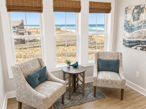 A room with two patterned armchairs, a small round table with a plant, and a view of the beach through large windows with blinds.