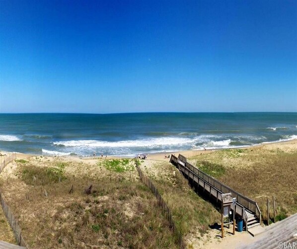 Ein Strand mit einer Holzpromenade, die zu einer kleinen Struktur, vermutlich einer Toilette oder Schutzhütte, am Wasserrand führt.