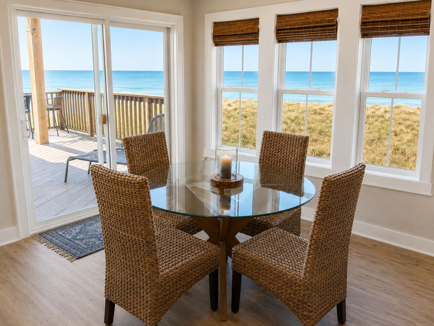 Une table ronde en verre, quatre chaises en osier, une bougie et une vue sur la plage à travers de grandes fenêtres.