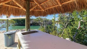 A beachside lounge with a thatched roof, a wooden bench, and a view of the sea and greenery. - Dbl Master |Oceanview |Jacuzzi |Beach |Marina |BBQ (Tavernier)
