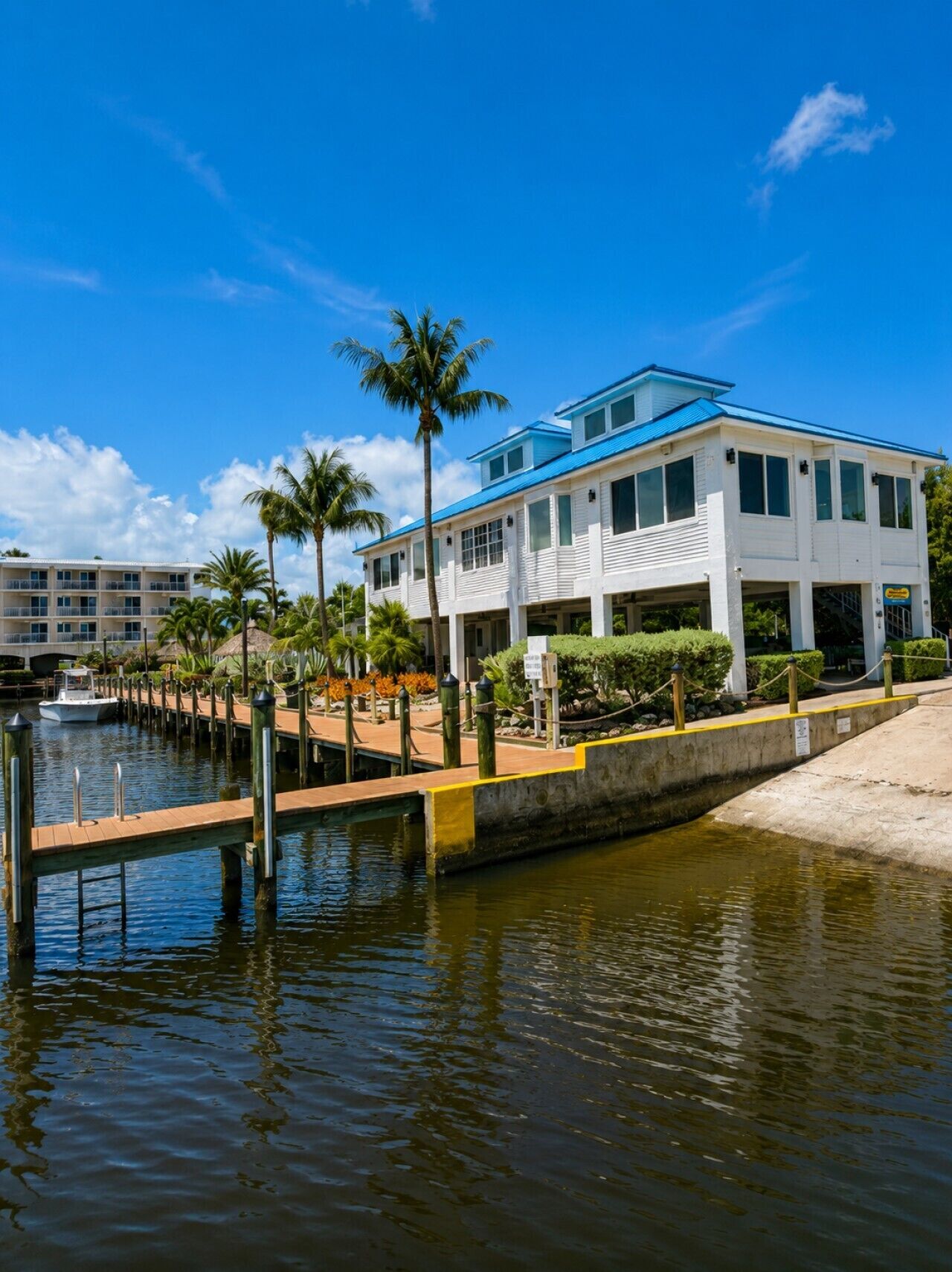 Hotel aan het water met een steiger, palmbomen en een helderblauwe lucht.