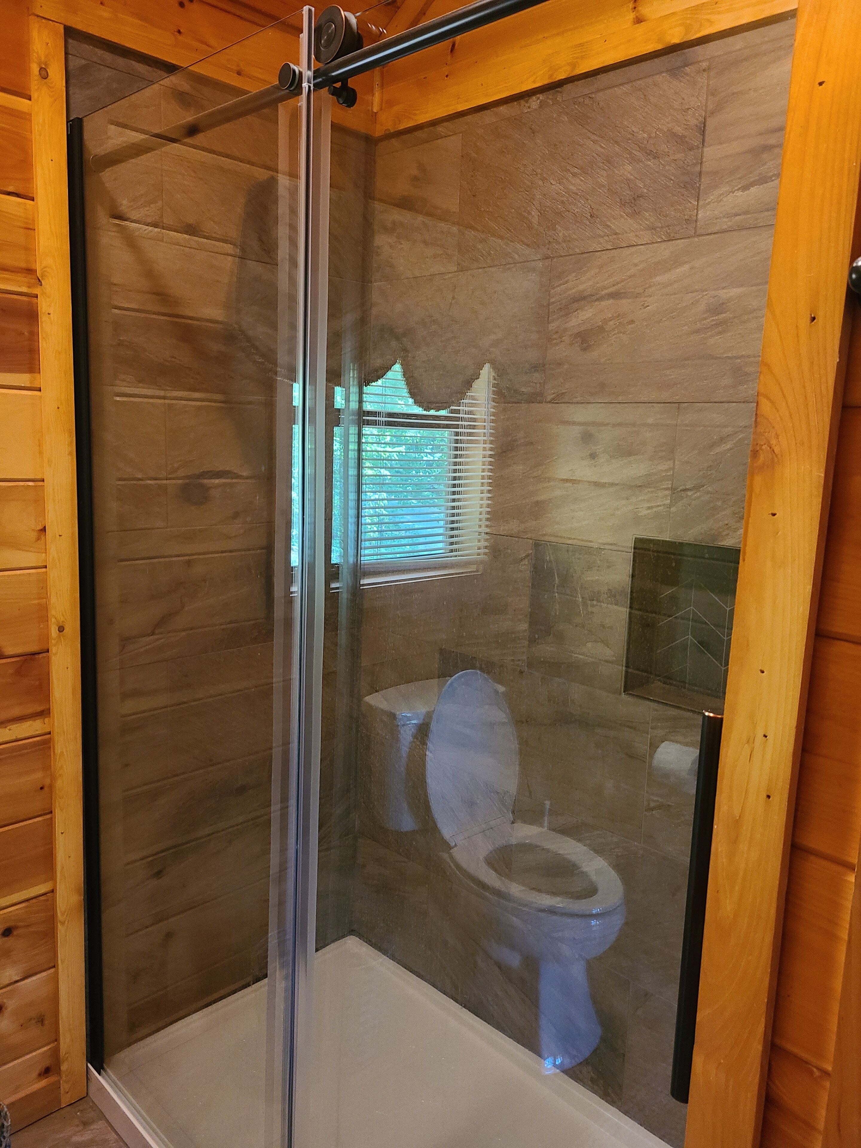 A glass-enclosed shower stall with a wooden interior, a toilet visible through the glass, and a window with blinds.