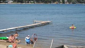 A lakeside scene with a wooden dock extending into the water, a sandy beach, and a few people and boats. - Sand Lake Cottage - near Tawas (National City)