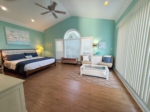A bedroom with a bed, a ceiling fan, a window with blinds, a chair, a dresser, and a lamp.