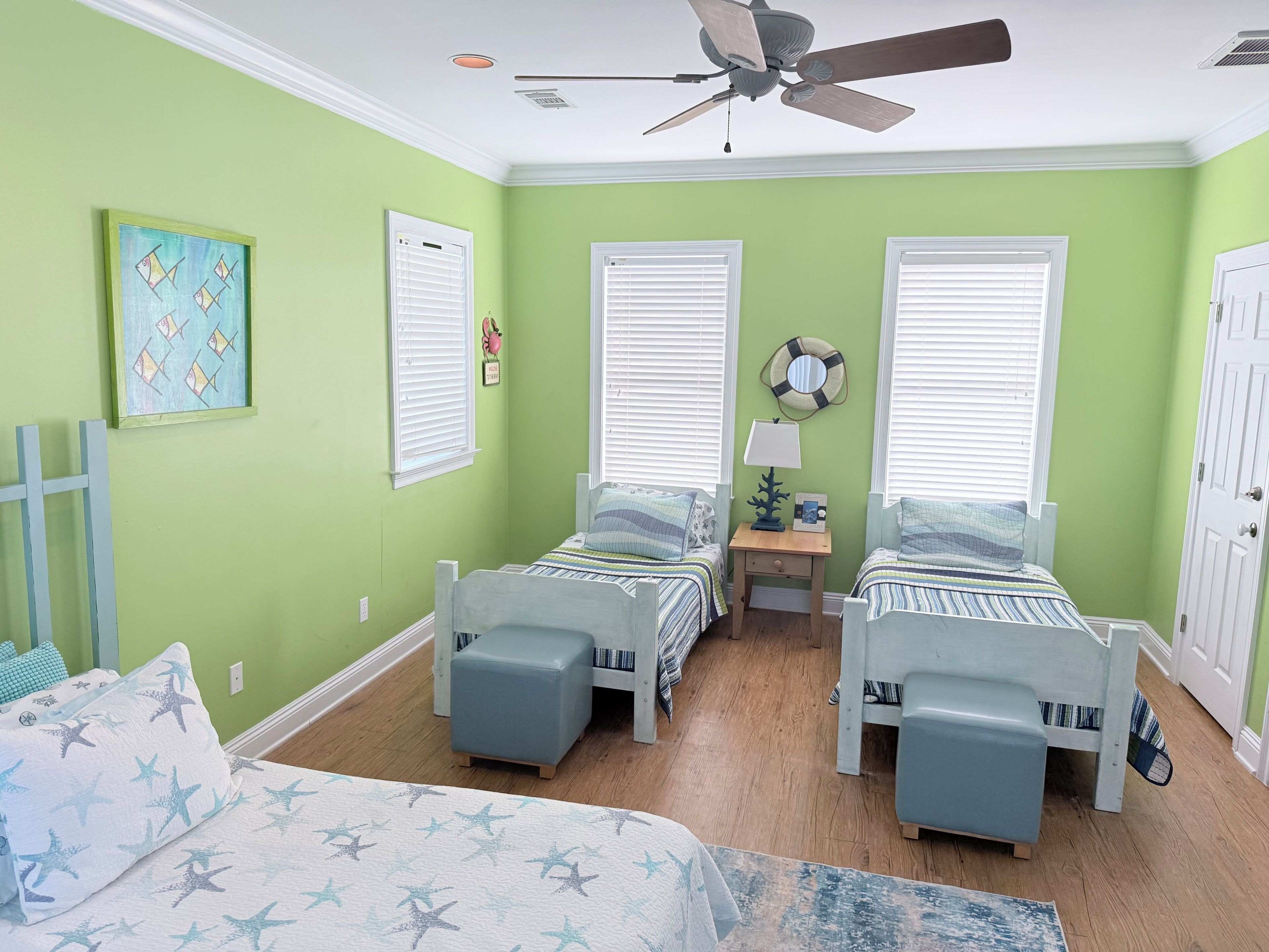 A room with two beds, a ceiling fan, a small table with a lamp, and a wall with a framed picture.