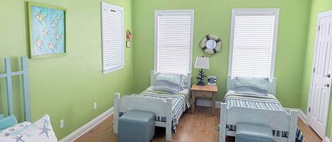 A room with two beds, a ceiling fan, a small table with a lamp, and a wall with a framed picture.
