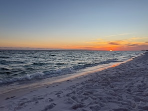 Una playa con huellas que van hacia el agua, un atardecer de fondo y un mar tranquilo.