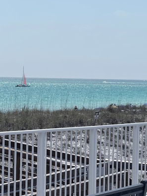 Vista costera con un velero en el agua, una playa de vegetación escasa y una cerca blanca al frente.