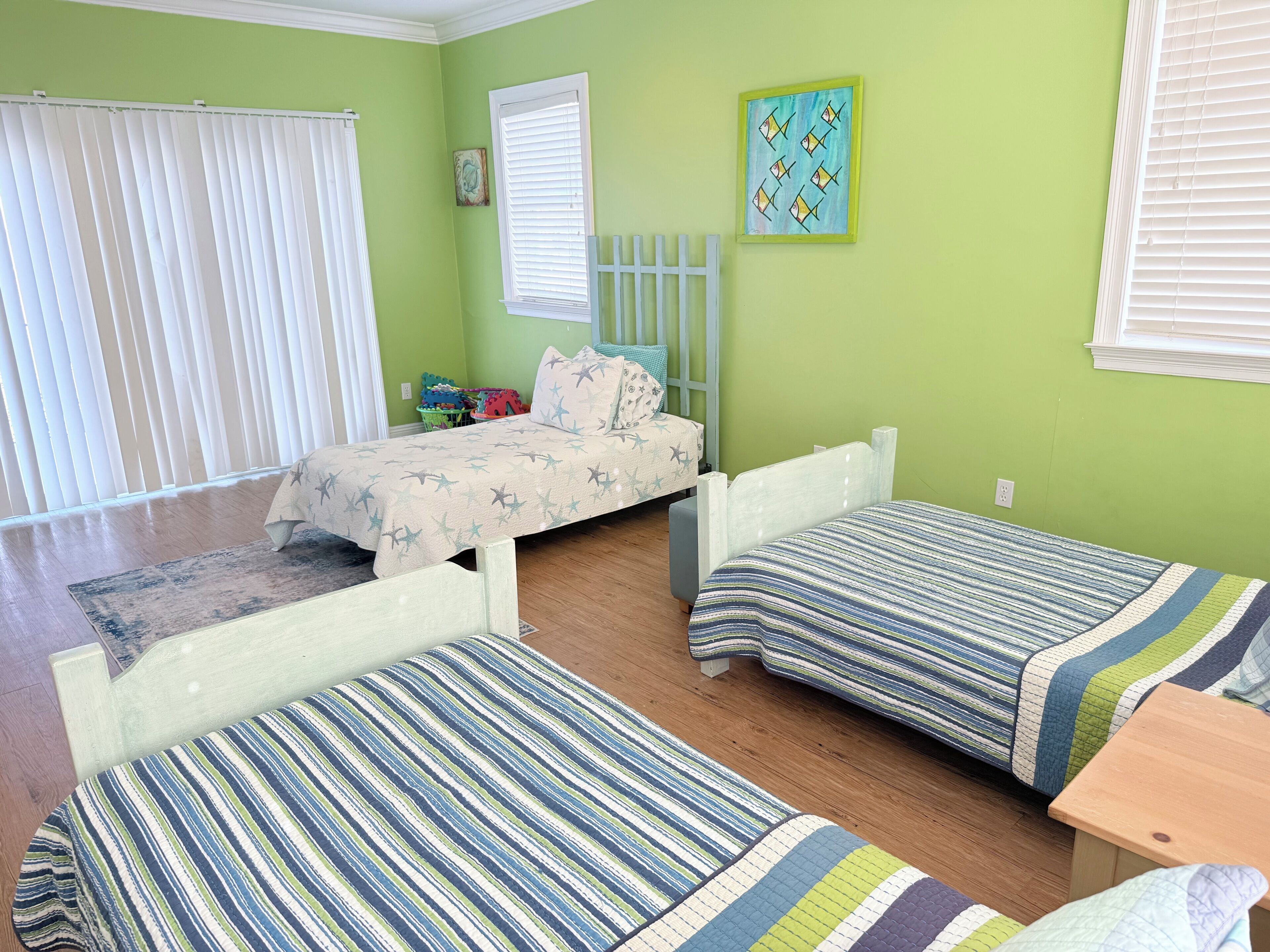 A room with two beds, a window with blinds, a small table, and a colorful painting on the wall.