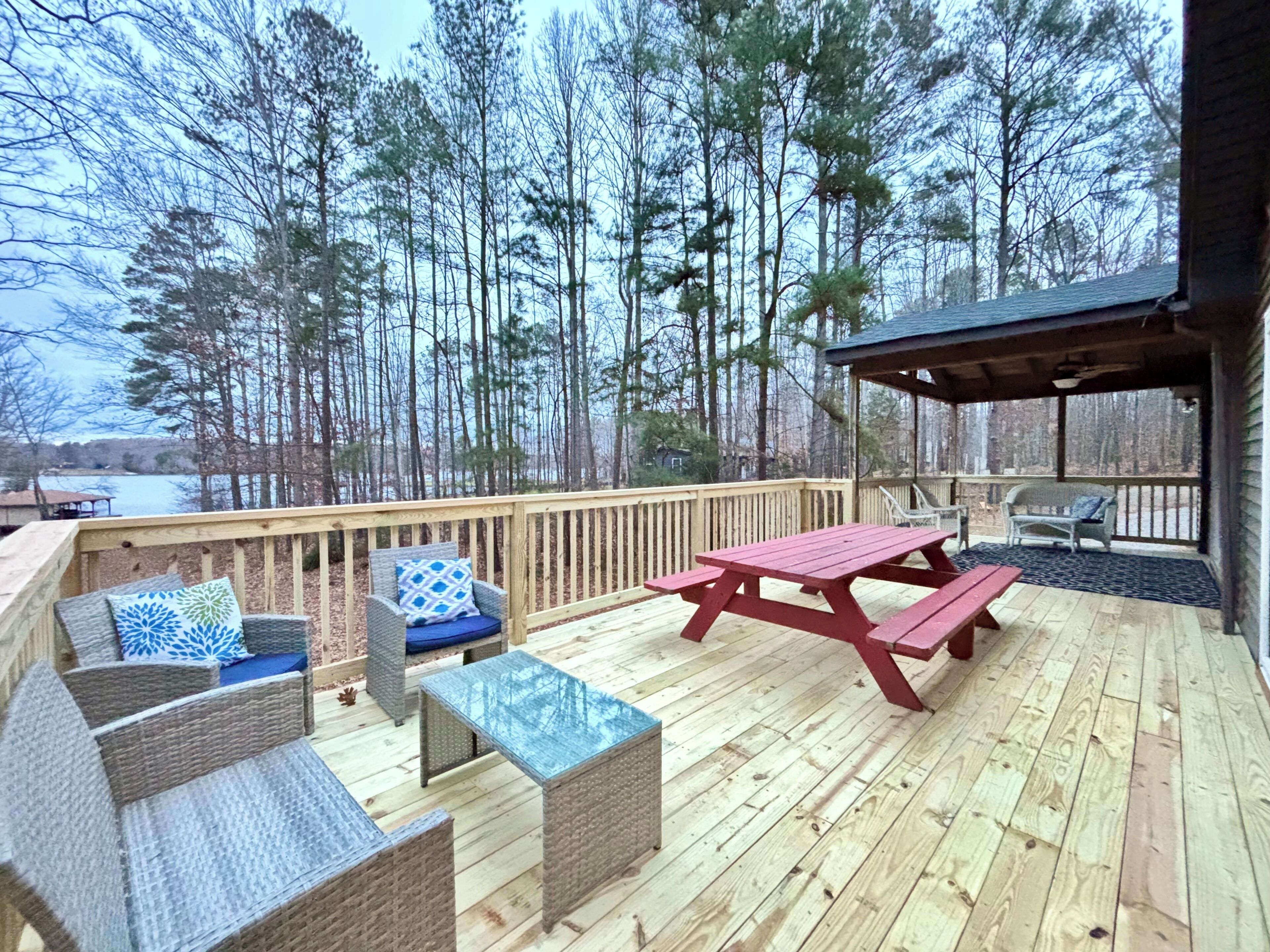 Una terraza de madera con muebles de mimbre, una mesa de picnic y una zona de estar cubierta con vistas a un bosque y una masa de agua.