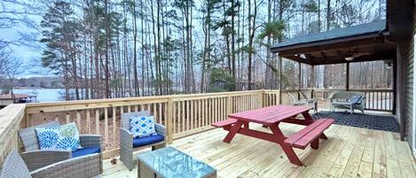Una terraza de madera con muebles de mimbre, una mesa de picnic y una zona de estar cubierta con vistas a un bosque y una masa de agua.