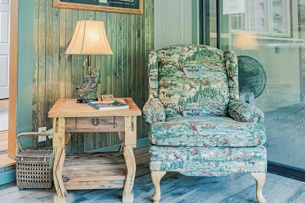 Una habitación con un escritorio de madera, un sillón estampado, una lámpara y un ventilador.