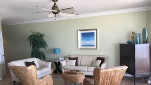 A living room with a ceiling fan, a sofa, two wicker chairs, a coffee table, a lamp, a framed picture, a plant, and a cabinet.