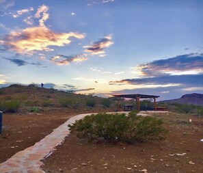 Paisaje desértico con un camino pavimentado que conduce a una construcción con techo, rodeada de vegetación escasa.