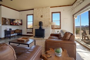 A living room with a stone fireplace, leather sofas, a desk, and a large window with a view of the desert. - Crystal Valley Cabin: Modern-Rustic Style&Comfort (Alpine)