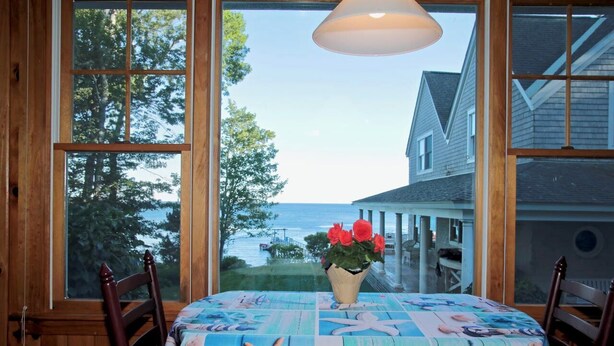 A dining table with a nautical-themed tablecloth, a vase with red flowers, and a view of a body of water and houses through large windows. - Come and enjoy our Boothbay Cottage (Boothbay Harbor)