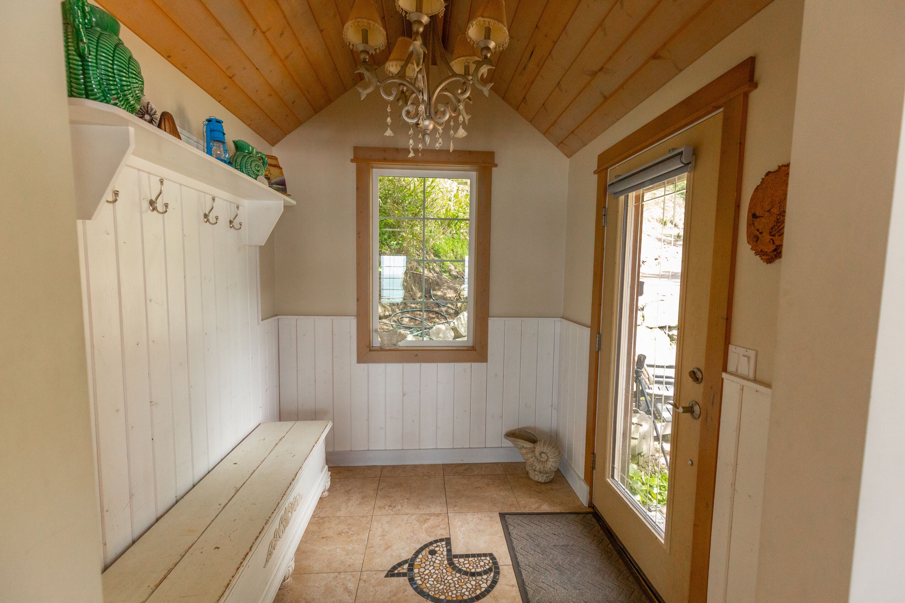 A narrow hallway with a wooden ceiling, a window with a view of greenery, a door with a glass panel, and a bench with hooks on the wall.