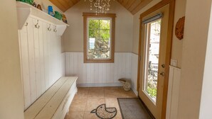 Un pasillo estrecho con techo de madera, una ventana con vistas a la vegetación, una puerta con panel de vidrio y un banco con ganchos en la pared.